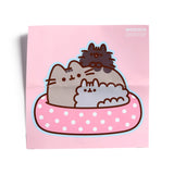 Front view of a pink wooden jigsaw puzzle depicting a grey Pusheen cat and two smaller cats snuggled in a pink polka dot bed.
