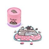 Angled view of a partially assembled 130-piece wooden Pusheen the Cat jigsaw puzzle, showing grey Pusheen cats on a pink polka-dot blanket, with loose pieces. A pink cylindrical storage tube with a Pusheen illustration is also visible on a white background.