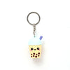 Front view of a 3D PVC bubble tea keyring. It features a light yellow cup with dark brown boba, a cute cartoon face with pink cheeks, a light blue lid, and a purple straw, attached to a metal ring and chain.