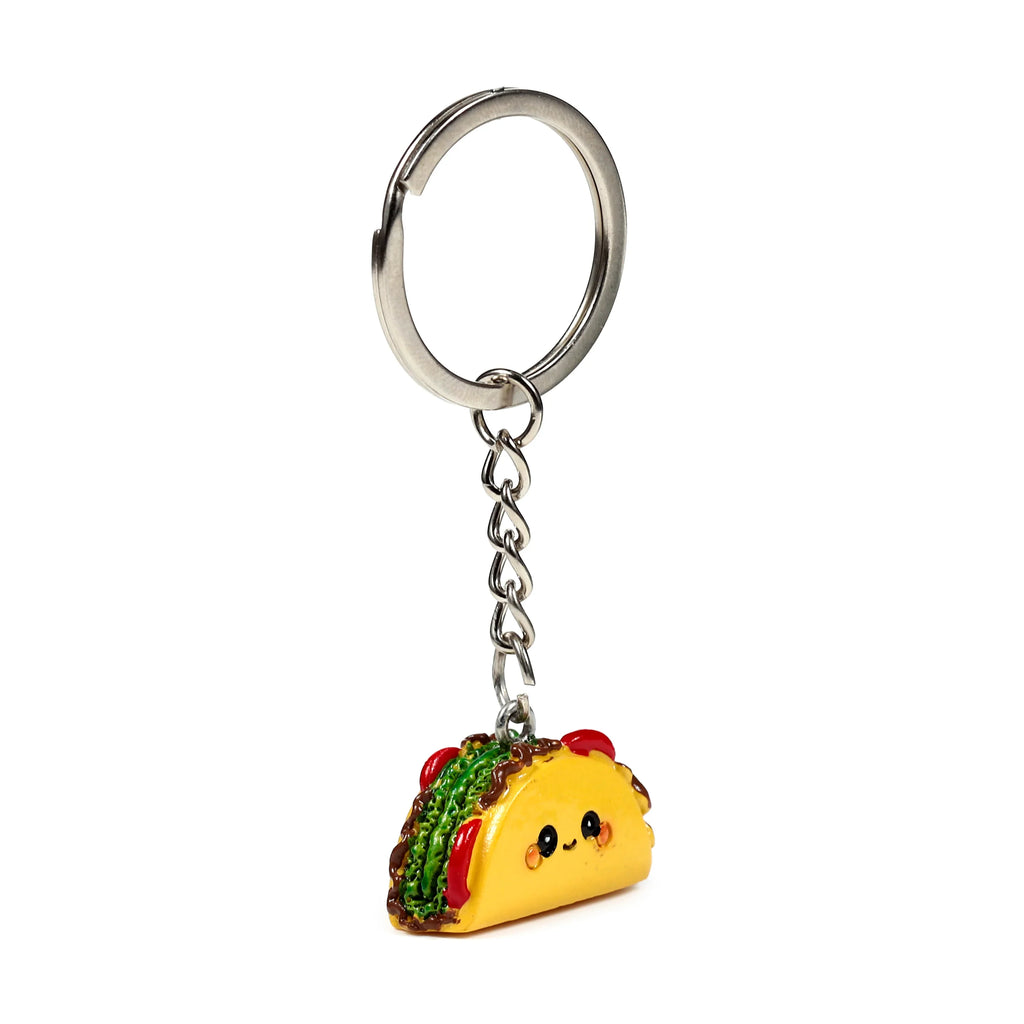 Angled view of a novelty keyring featuring a yellow taco charm with a smiling face, green, red, and brown fillings, attached to a silver-coloured metal split ring and chain.
