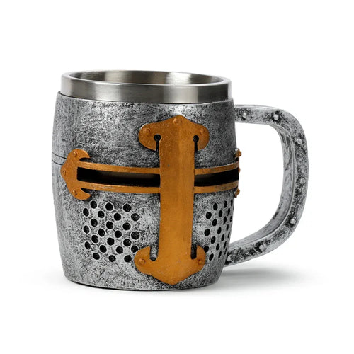 Front view of a decorative medieval knight helmet tankard, crafted with a textured silver-effect finish. It features a prominent gold-coloured cross design and black perforated details mimicking a helmet visor. The tankard has a metallic interior rim and a robust handle.