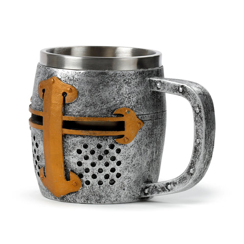 Angled view of a decorative silver-coloured medieval knight helmet tankard with a distressed metallic finish, featuring a gold-coloured cross design and perforated visor section on the front, and a textured handle.