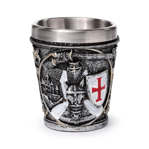 Front view of a decorative shot glass with a textured grey exterior and silver-coloured inner, featuring a medieval knight in armour holding a white shield with a red cross, surrounded by dragon and castle motifs within an ornate gothic-style design.