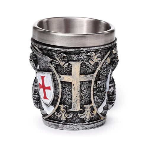 Angled view of a decorative medieval knight shot glass, showcasing a central raised aged gold-coloured cross and a white shield with a red cross, against an intricately textured metallic grey and silver background.