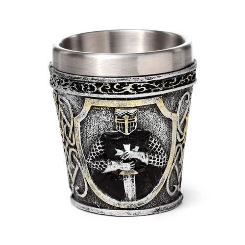 Front view of a decorative medieval shot glass, featuring a textured silver-grey exterior with a raised design of a black knight in armour. The knight, holding a sword, has a white cross on his chest, set within a gold-bordered shield, surrounded by intricate patterns.