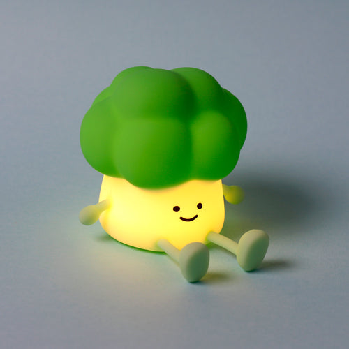 Angled view of a cute green broccoli character night light, glowing yellow, with a happy face, sitting on a pale blue background.