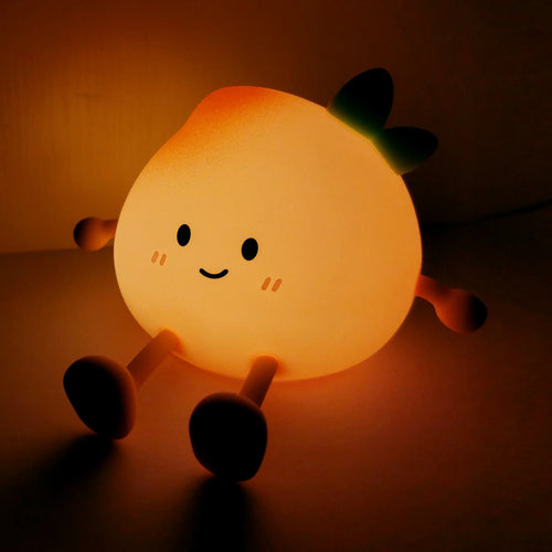 Angled view of an illuminated peach character night light, featuring a smiling cartoon face, black eyes, and green leaves on its top, sitting and emitting a warm orange glow.