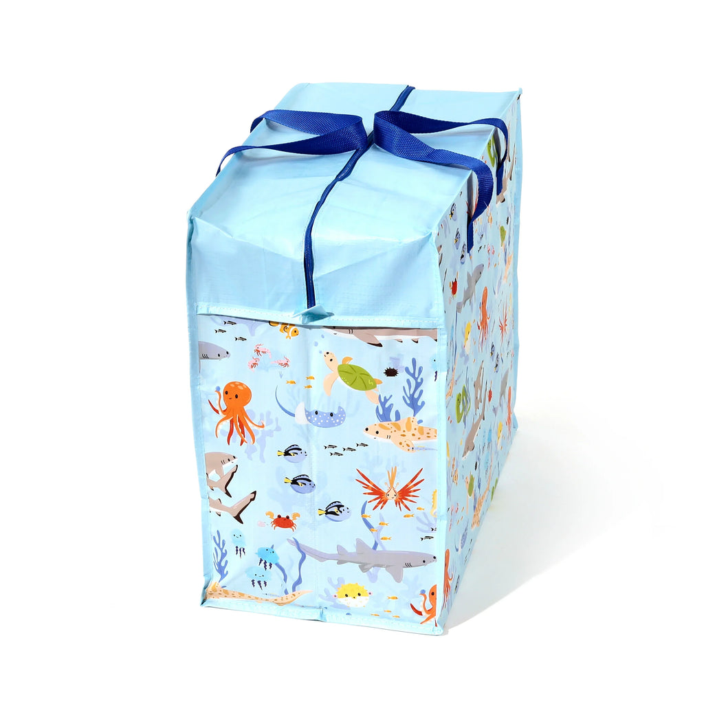 Angled view of a light blue Mariniverse laundry and storage bag, decorated with a colourful pattern of cartoon sea creatures including octopuses, sharks, turtles, and fish. The bag features a zippered light blue lid and dark blue fabric handles tied in a bow.