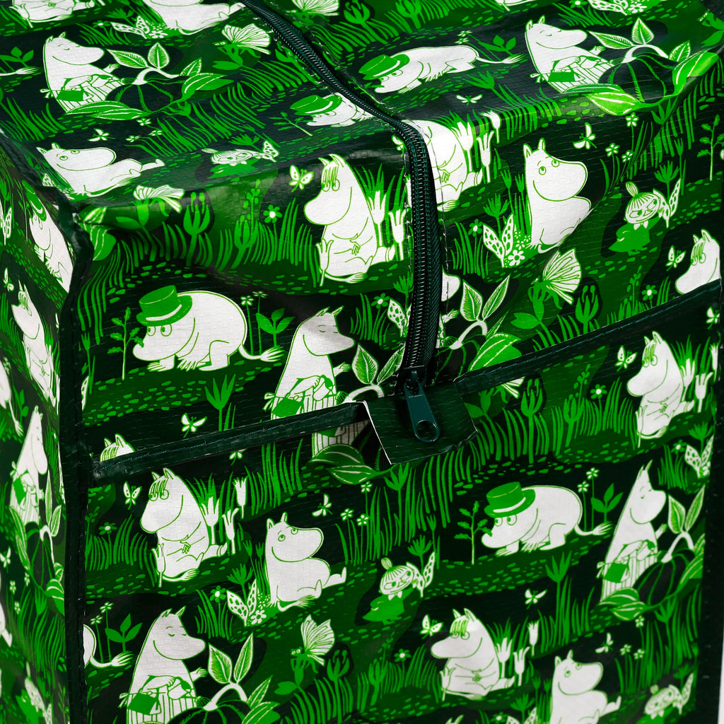 Close-up, angled view of a green laundry and storage bag with a white Moomin garden life pattern and a dark green zip.