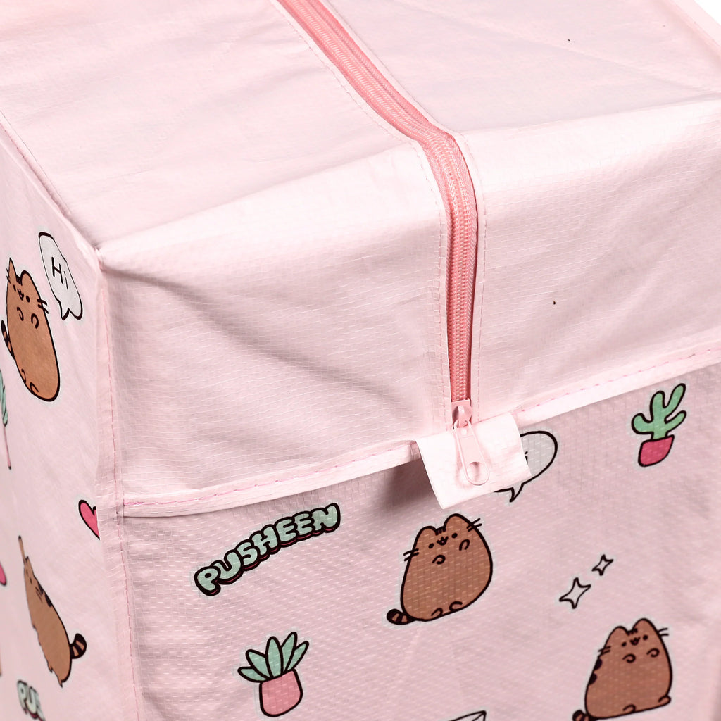 Close-up, angled view of a pink Pusheen the Cat laundry and storage bag, showing the top zip closure with a pink zipper pull and part of the side panel. The light pink textured fabric features a repeat pattern of Pusheen cats, some with speech bubbles, the word 'PUSHEEN', and small potted plant illustrations.