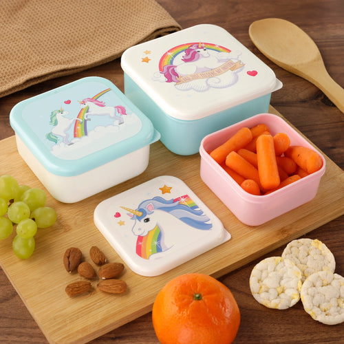 In-context lifestyle shot of a set of three colourful rainbow unicorn lunch boxes (S, M, L) on a wooden chopping board. The lunch boxes feature varied unicorn designs, with the smallest pink-based box open and containing carrots. Grapes, almonds, rice cakes, and an orange are also visible.