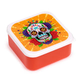 Angled view of a square, reddish-orange lunch box with a white-rimmed lid, featuring a vibrant Day of the Dead sugar skull design. The skull is decorated with colourful floral and geometric patterns, set against an orange, speckled background.