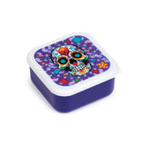 Angled view of a colourful Day of the Dead sugar skull lunch box. It has a deep purple base and a white lid, which features an ornate multi-coloured skull design amidst patterns of diamonds, flowers, hearts, and confetti.