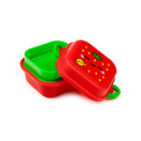 Angled view of a partially open red and green bento clip lock snack box. The red lid features a cartoon strawberry design with black eyes, pink cheeks, yellow seeds, and a yellow 'FRESH' star, revealing a bright green inner compartment and a green clip lock.