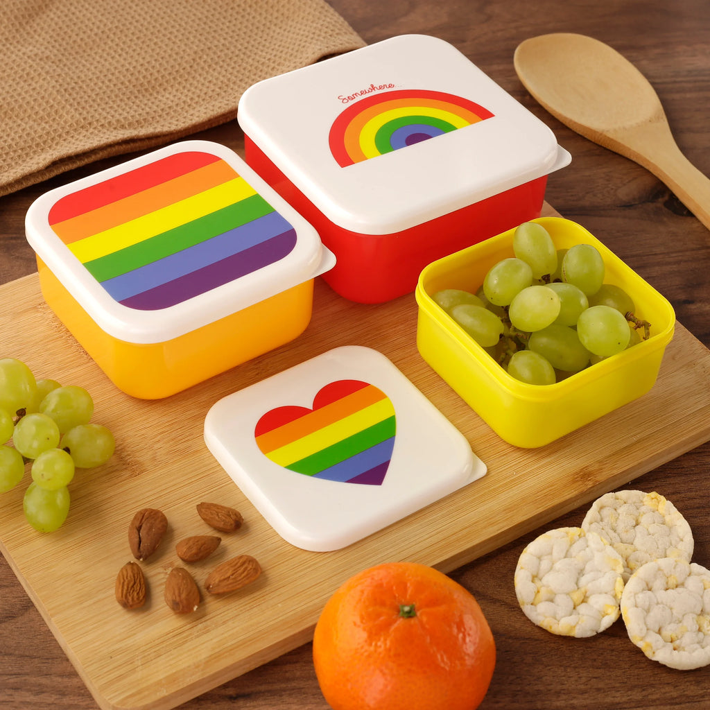 Overhead lifestyle shot of a set of three rainbow lunch boxes, featuring white lids with an arc rainbow, striped rainbow, and a rainbow heart design. The lunch boxes have one red and two yellow bases, and are arranged on a wooden board with grapes, almonds, a tangerine, and rice cakes.