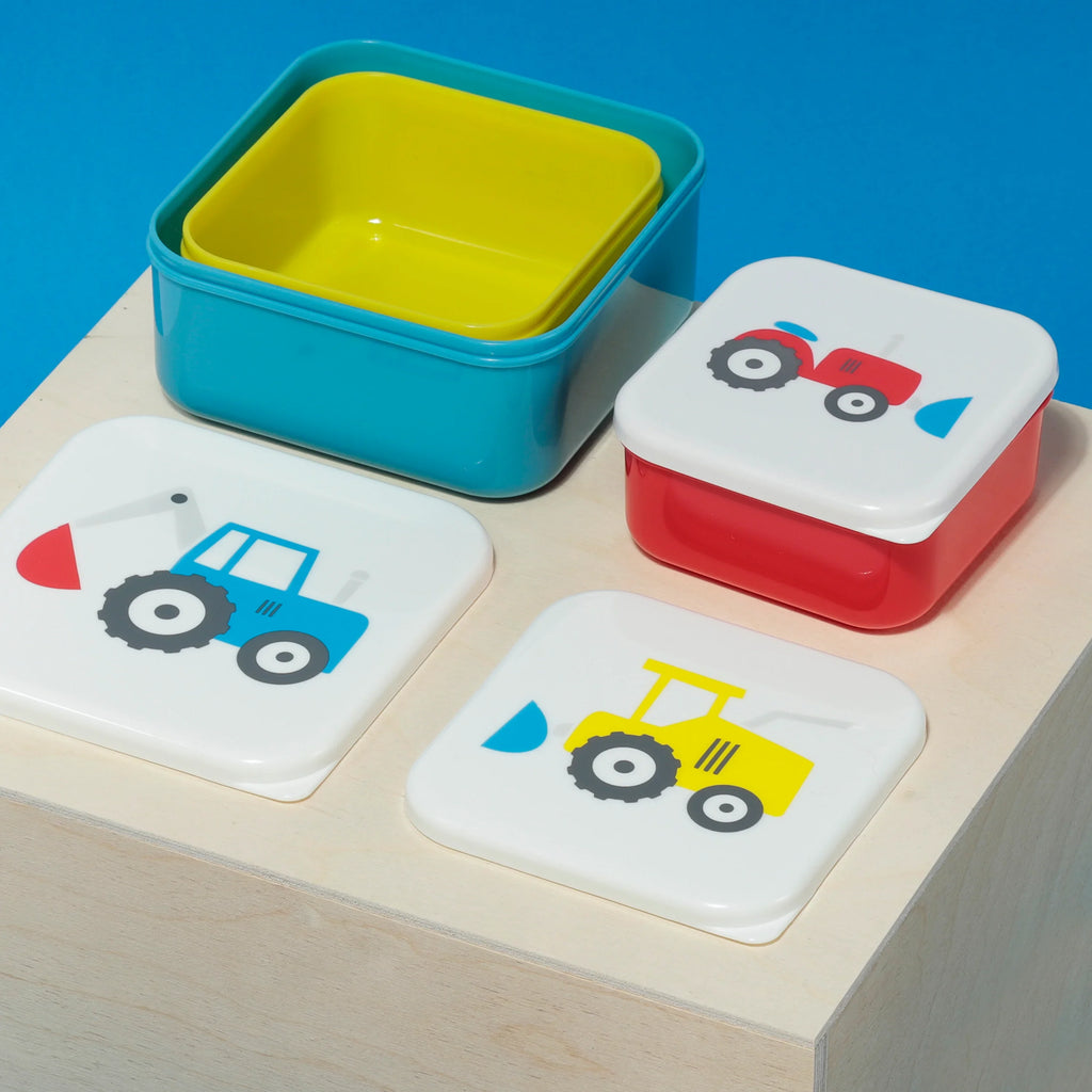 Angled top-down view of a bright turquoise blue lunch box base with a yellow inner compartment. Alongside it are a red lunch box with its white lid depicting a red tractor, and two additional white lids showing a blue digger and a yellow tractor design. All items are arranged on a light wooden surface against a blue background.