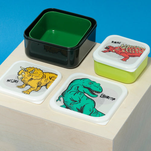 Angled view from above of a set of three colourful dinosaur lunch boxes. The largest dark green box nests a bright green one; their separate white lids display a teal T-Rex with 'CHOMP' text and a yellow Triceratops with 'grRR!' text. A third, smaller lime green lunch box is assembled, its white lid featuring a red Ankylosaurus with 'ROAR!' text.