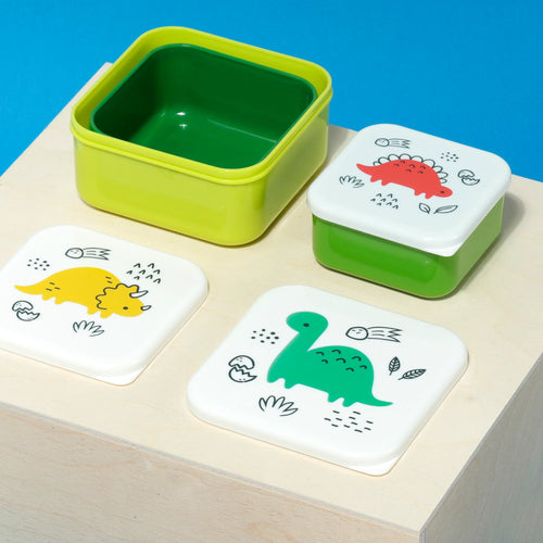 Overhead angled view of five colourful lunch boxes on a wooden surface. A large lime green box contains a dark green nesting box. Three smaller white-lidded boxes are also visible, featuring red, yellow, and green cartoon dinosaur designs.