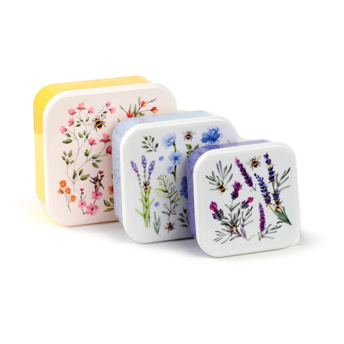Angled view of three lunch boxes, from largest to smallest, showcasing their patterned lids and contrasting bases. The largest lunch box features a cream lid with pink and orange floral designs and bees, and a yellow base. The medium has a white lid with blue floral designs and bees, and a light blue base. The smallest displays a white lid with purple lavender designs and bees, and a light purple base. All are presented on a clean white background.