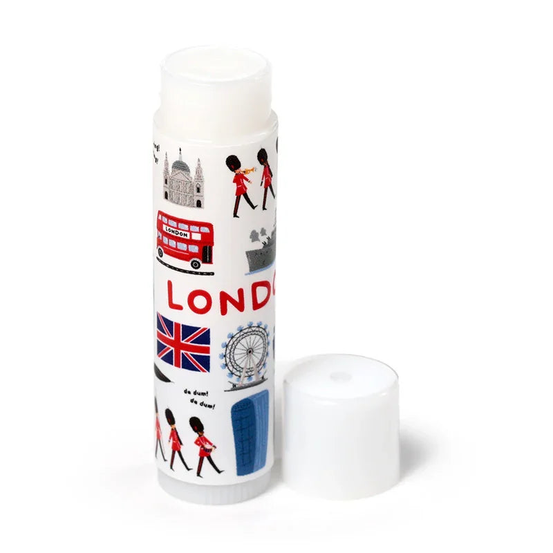London souvenir stick lip balm, white, standing upright with cap removed. Features illustrations of a red double-decker bus, Union Jack flag, London Eye, Queen's Guards, and the word 'LONDON'.