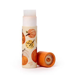 Angled view of an autumn harvest paper stick lip balm, showing the cream-coloured tube with various pumpkin illustrations. The white balm is extended from the top, and the orange cap with a small pumpkin design rests on its side beside it.