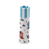 Angled view of a cylindrical coastal-themed paper stick lip balm. It features a light blue and white striped lid, and the white body is illustrated with beach huts, a sailboat, sunglasses, flip-flops, and a seagull.