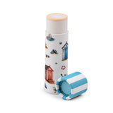 Angled view of an upright Paper Stick Lip Balm showing its natural-coloured balm and a white tube with coastal beach illustrations including beach huts and a sailboat. Its detached blue and white striped cap, marked Plastic Free, 100% recyclable, lies beside it.