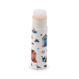 Upright paper stick lip balm with translucent white balm and a peachy layer partially extruded, featuring a white tube patterned with colourful beach huts, a sailing boat, flip-flops, sunglasses, and a seagull.