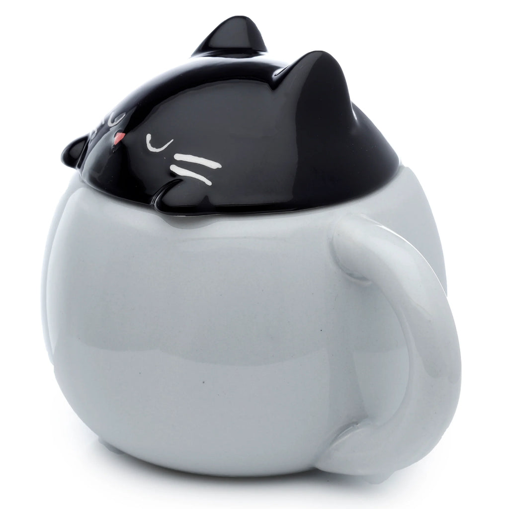 Angled view of a light grey ceramic lidded animal mug, with a black cat's head lid featuring pointed ears, closed white eyes, pink nose, and white whiskers, handle visible.