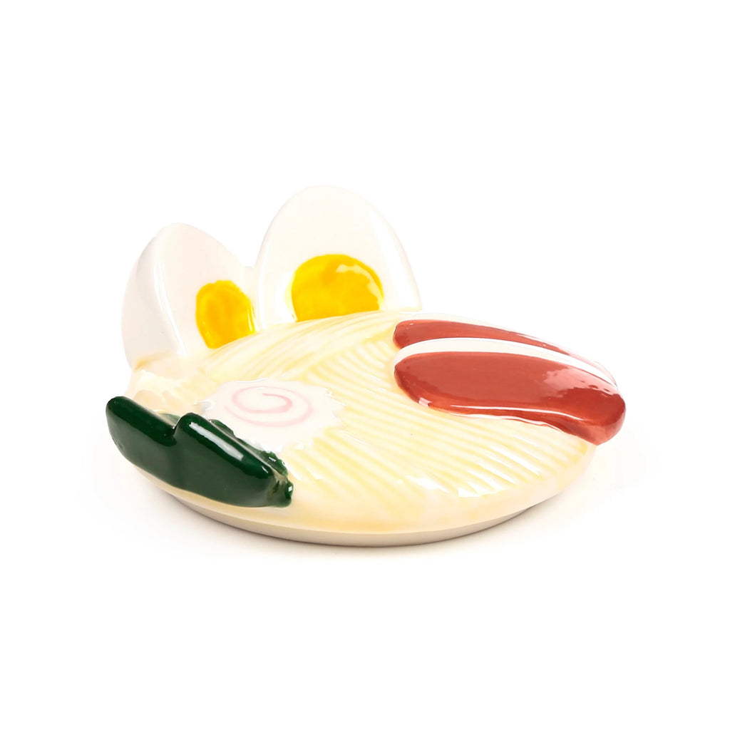 Angled view of a ceramic ramen mug lid, sculpted with noodles, egg halves, narutomaki, and green garnish.