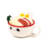 Angled view of a white ceramic lidded mug with a smiling face, a visible handle, and a ramen noodle lid decorated with eggs, char siu, and spring onions.