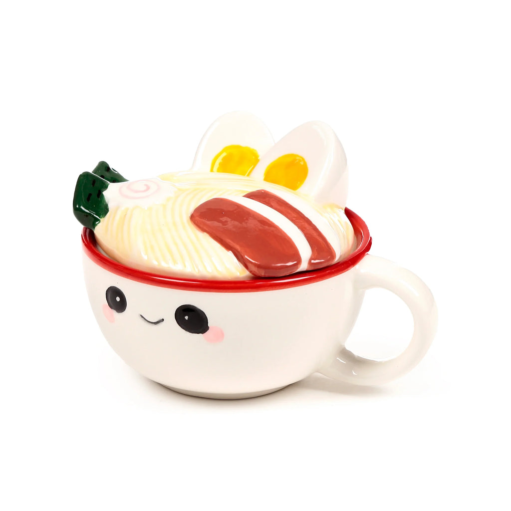 Angled view of a white ceramic lidded mug with a smiling face, a visible handle, and a ramen noodle lid decorated with eggs, char siu, and spring onions.