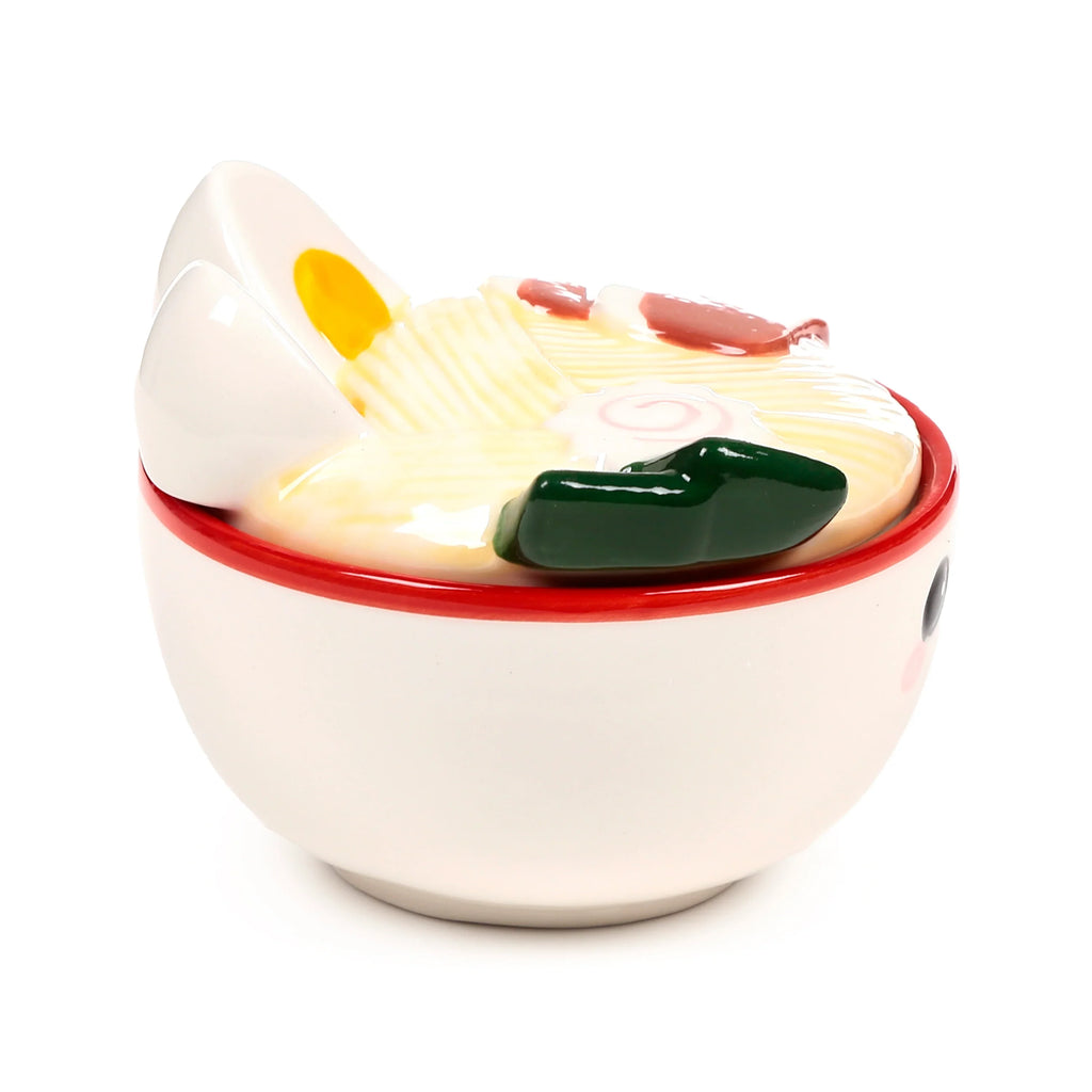 Angled view of a cream ceramic lidded mug with a red rim and cute face. The lid is shaped like a ramen bowl, depicting an egg slice, yellow noodles, pink swirled fish cake, and dark green seaweed.