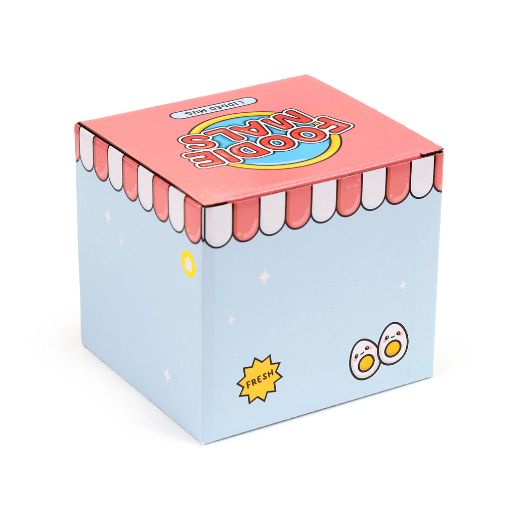 Angled view of the Ceramic Lidded Mug box, featuring pale blue sides with cartoon eggs and a 'Fresh' label, and a pink top with 'Foodiemals' text.