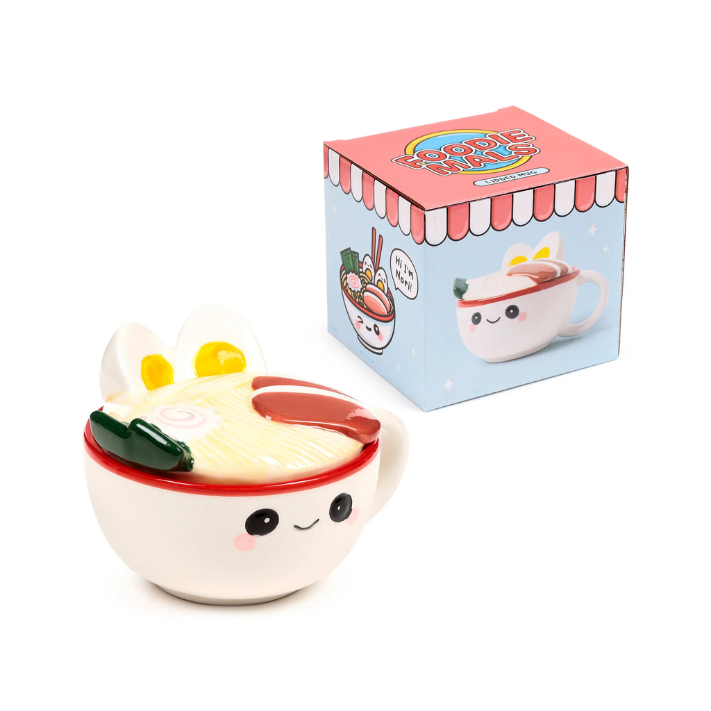 Angled view of a white ceramic Nori the Ramen Foodiemals lidded mug, depicting a smiling face on the mug and a ramen noodle lid with eggs, narutomaki, and seaweed. A light blue matching packaging box with cartoon ramen illustrations is visible in the background.