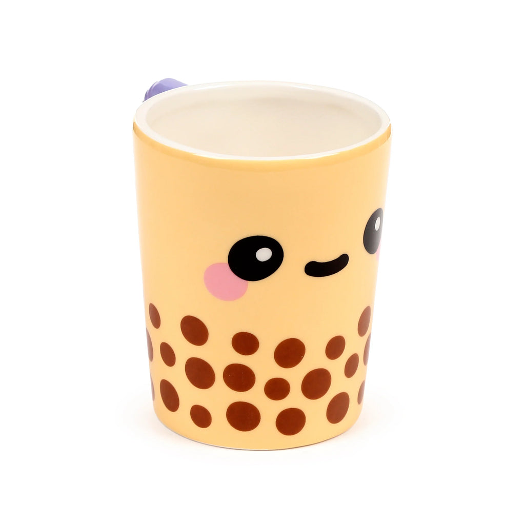 Angled view of a light beige ceramic mug, featuring a cute boba tea character design with black eyes, pink cheek blush, a small mouth, and brown circular 'pearl' patterns on its body. A small purple detail is visible at the top edge.