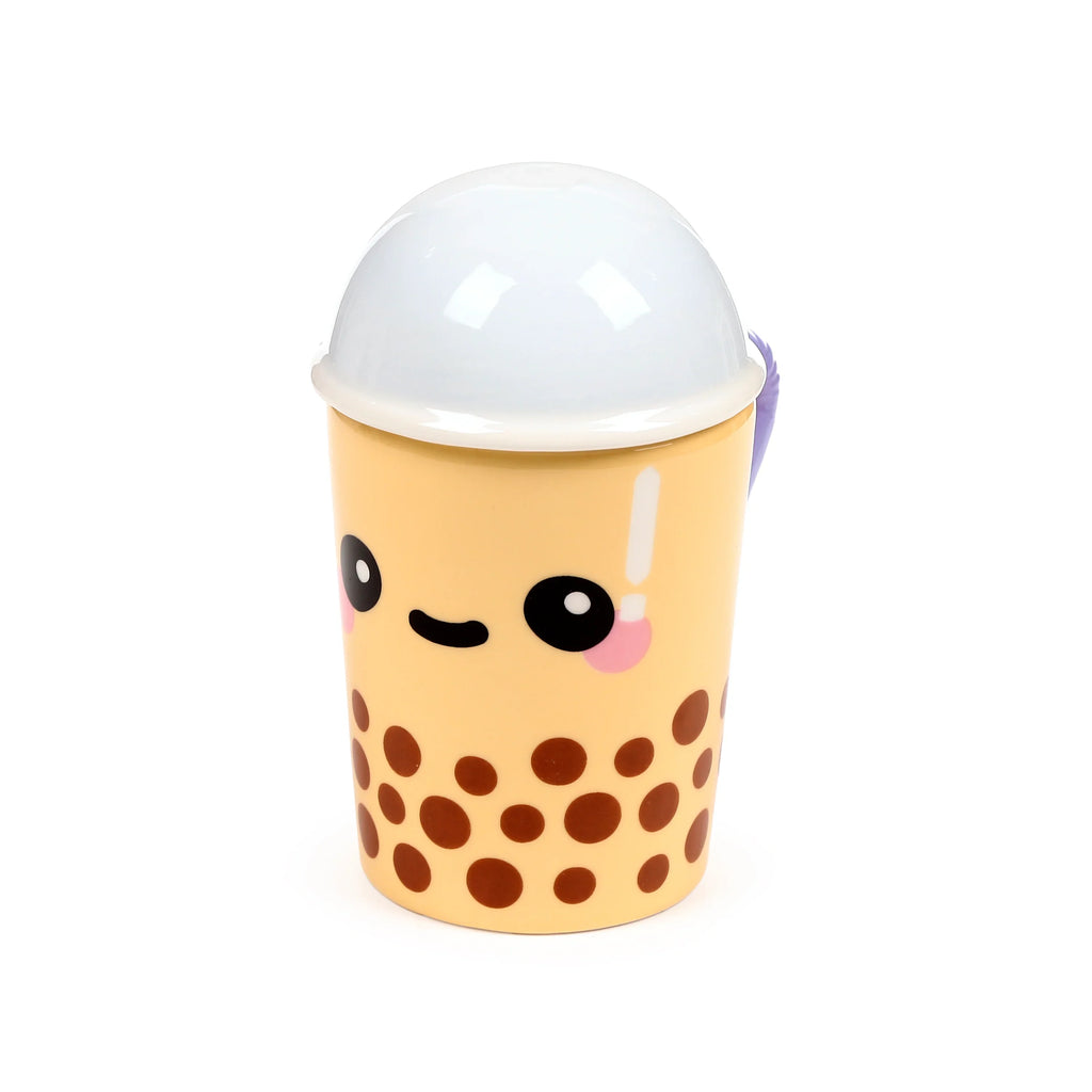 Angled view of a ceramic boba bubble tea lidded mug. The pale yellow mug features dark brown circular 'boba' patterns and a cute cartoon face with black eyes, a smiling mouth, and pink cheek blush. It is topped with a white domed lid, and a purple handle is partially visible on the right side.