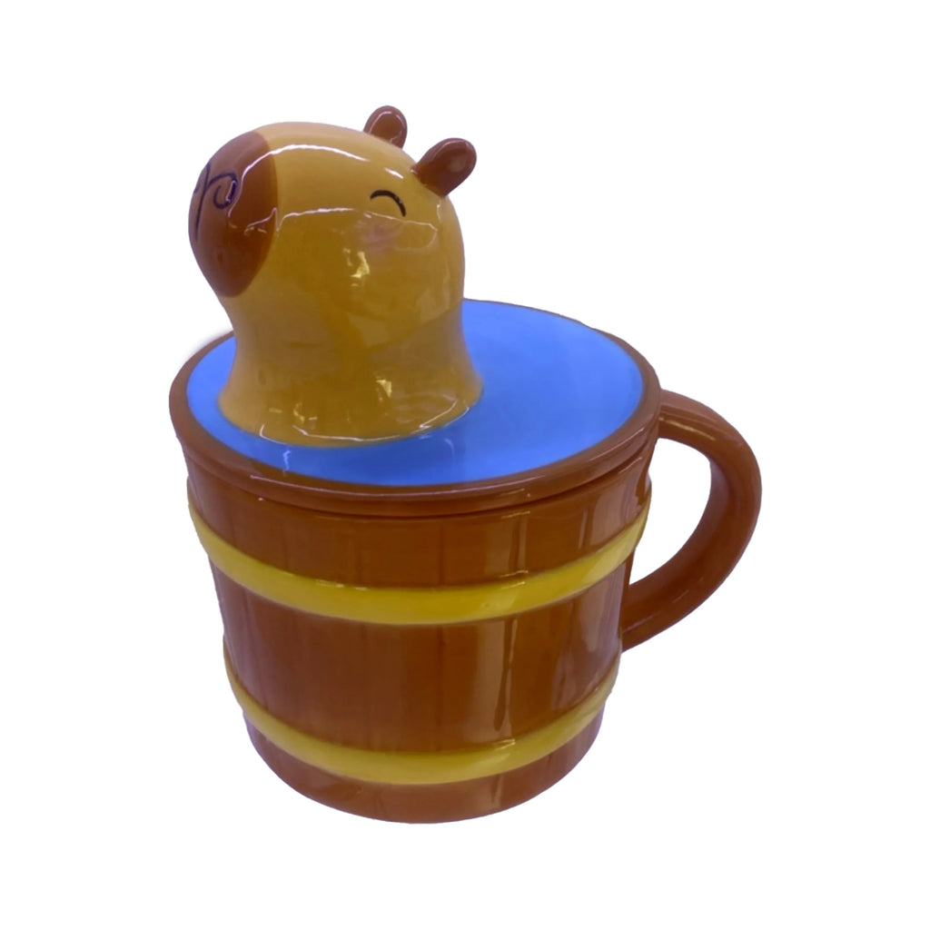 Elevated angled view of a capybara lidded ceramic mug. The brown barrel-shaped mug has yellow bands and a handle, with a smiling capybara head emerging from blue 'water' on the lid.