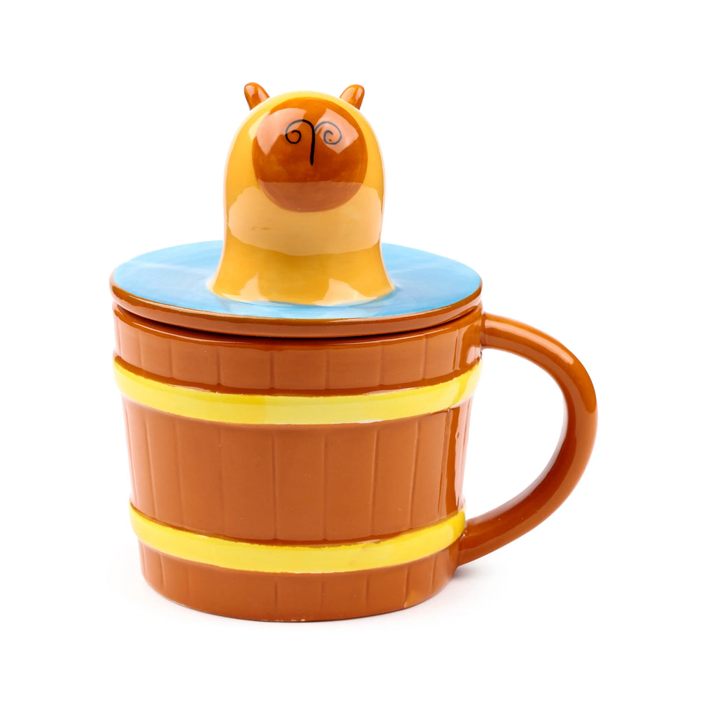 Ceramic lidded mug styled as a brown barrel with yellow bands, and a brown handle. The lid features a light blue top and a cartoon capybara head with dark brown features and small ears, facing the viewer and emerging from it.