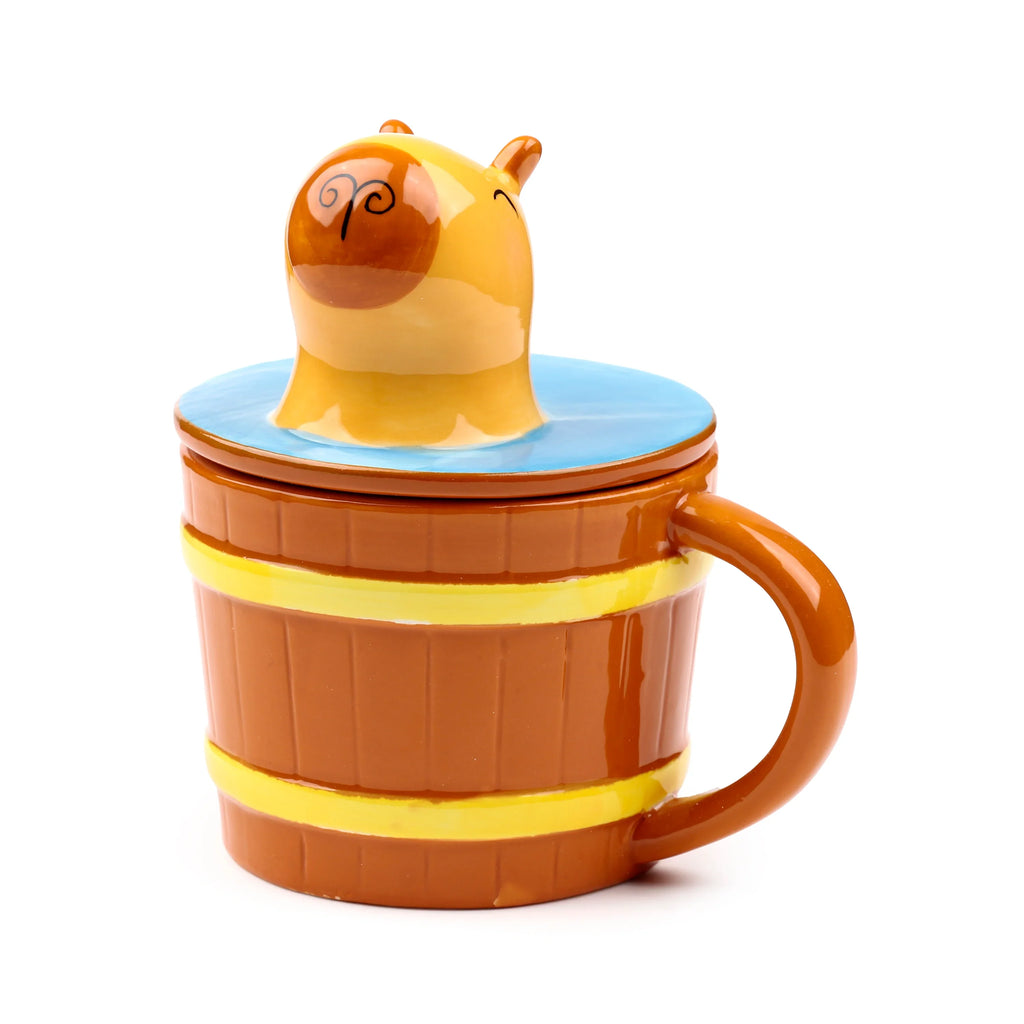 Angled view of a ceramic lidded mug, designed as a brown barrel with yellow bands, featuring a light brown capybara figure with small ears and darker brown markings emerging from a blue lid.