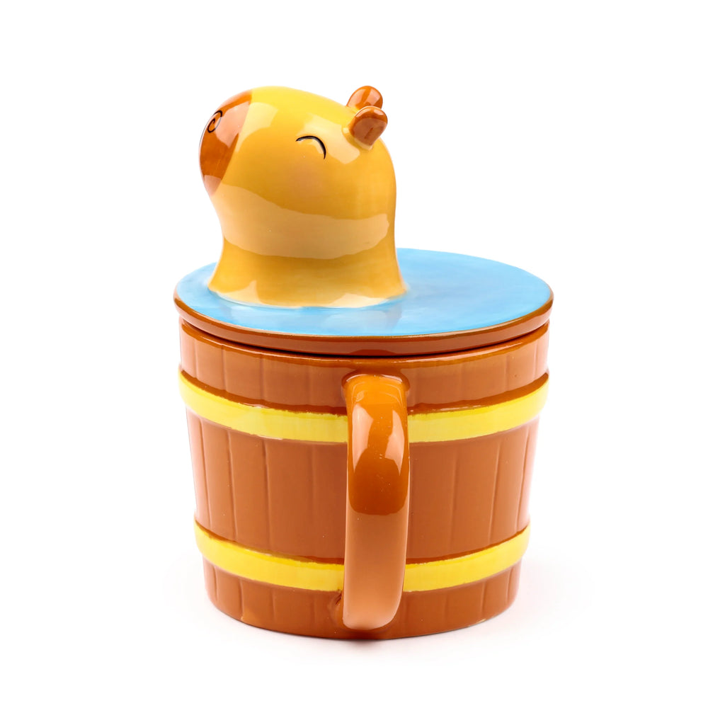Angled view of a ceramic capybara lidded mug, styled like a brown barrel with yellow bands. A capybara head, with closed eyes, emerges from blue on the lid, and a handle is visible on the side.