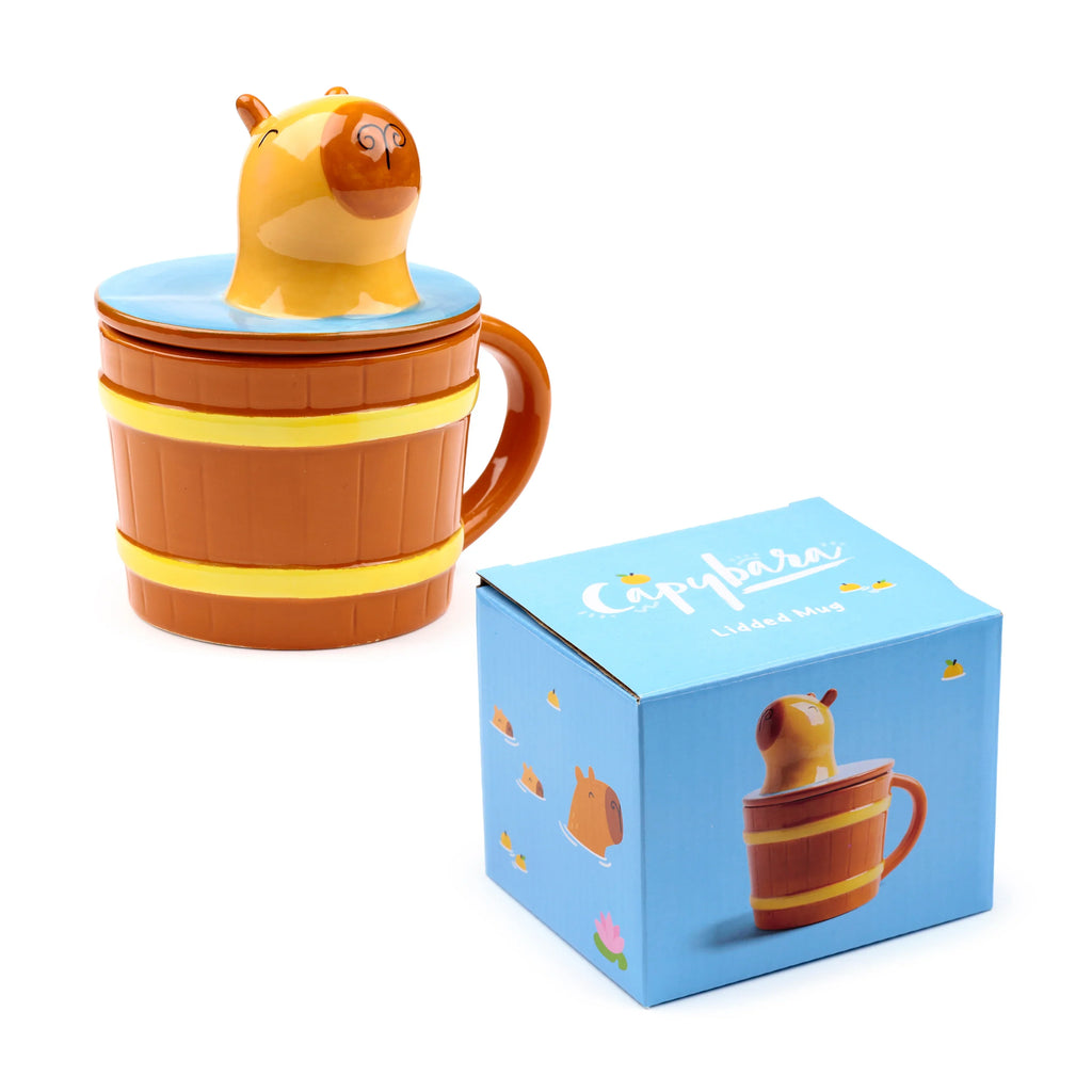 Angled view of a brown ceramic lidded mug, designed as a barrel with yellow bands, featuring a smiling capybara head emerging from its blue lid, next to its blue illustrated gift box.