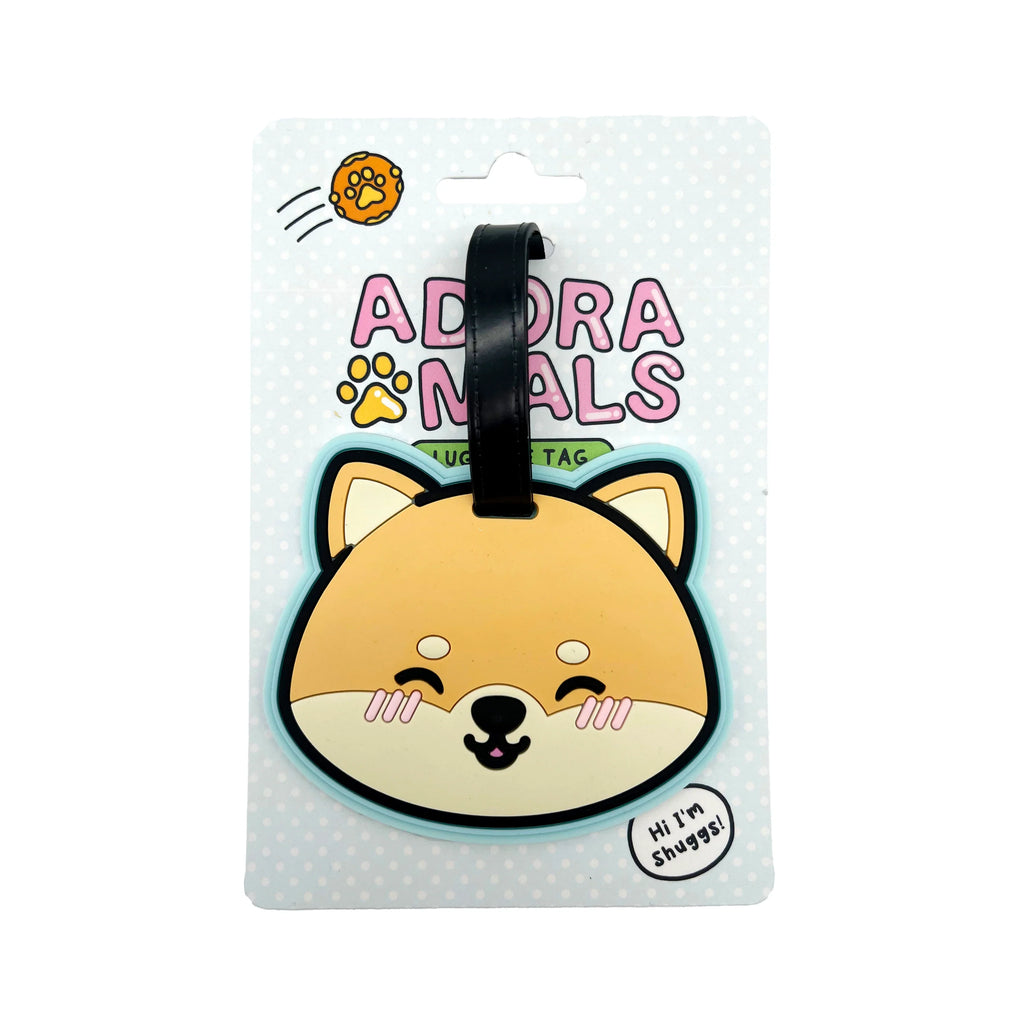 Front view of a PVC luggage tag shaped like a smiling cartoon Shiba Inu dog's head in tan, cream, black, and pink, with a light blue border and black strap, presented on light blue polka-dotted retail packaging.