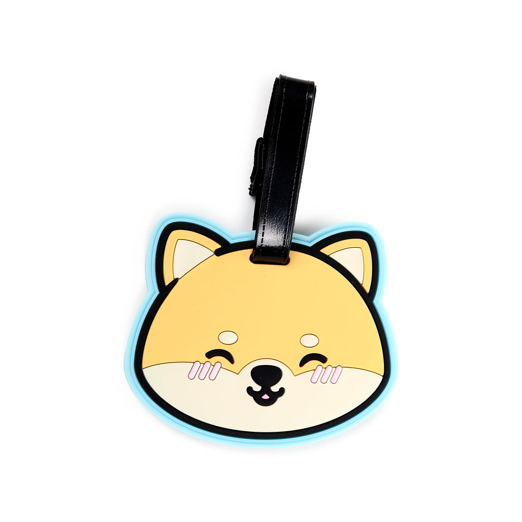 Front view of a PVC luggage tag shaped like a cartoon Shiba Inu dog's head, in beige and cream with black facial features, pink cheek blush, a light blue border, and a black strap.