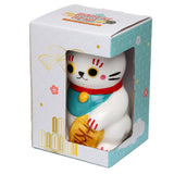 White ceramic Maneki Neko lucky cat money box, with red collar, turquoise bib, and gold coin, seen front-on through the window of its angled, patterned retail packaging.