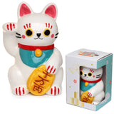 Front view of a white ceramic Maneki Neko lucky cat money box, with a raised paw and holding a gold coin, detailed with red, teal, black, and pink accents. It is displayed next to its decorative white and teal packaging box, which shows the cat through a cut-out window.