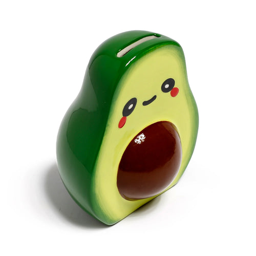 Angled view of a glossy ceramic avocado money box. It features a dark green skin, light green flesh with a cute cartoon face (black eyes, smile, red cheeks), a dark brown stone, and a coin slot on top.