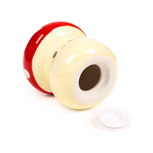 Angled view of a red and cream ceramic mushroom money box, lying on its side, with white spots on the cap and a cute face on the stalk. The coin slot opening is visible, and the white stopper is detached beside it.