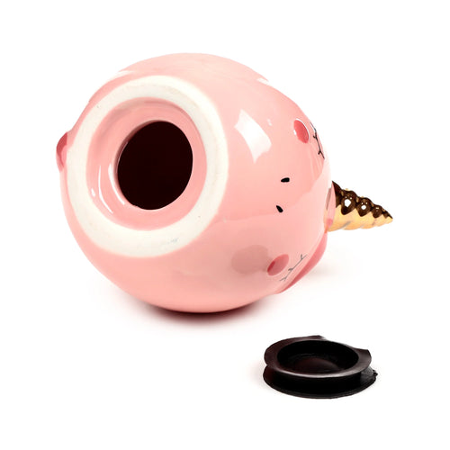 Angled view of a pink ceramic unicorn money box lying on its side, showing the coin retrieval opening at the base with a black rubber stopper, and a golden horn.