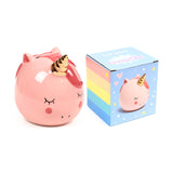 Angled view of a pink ceramic unicorn money box with a gold horn, closed eyes, and rosy cheeks, positioned beside its colourful rainbow-striped packaging box, which features a similar unicorn design.